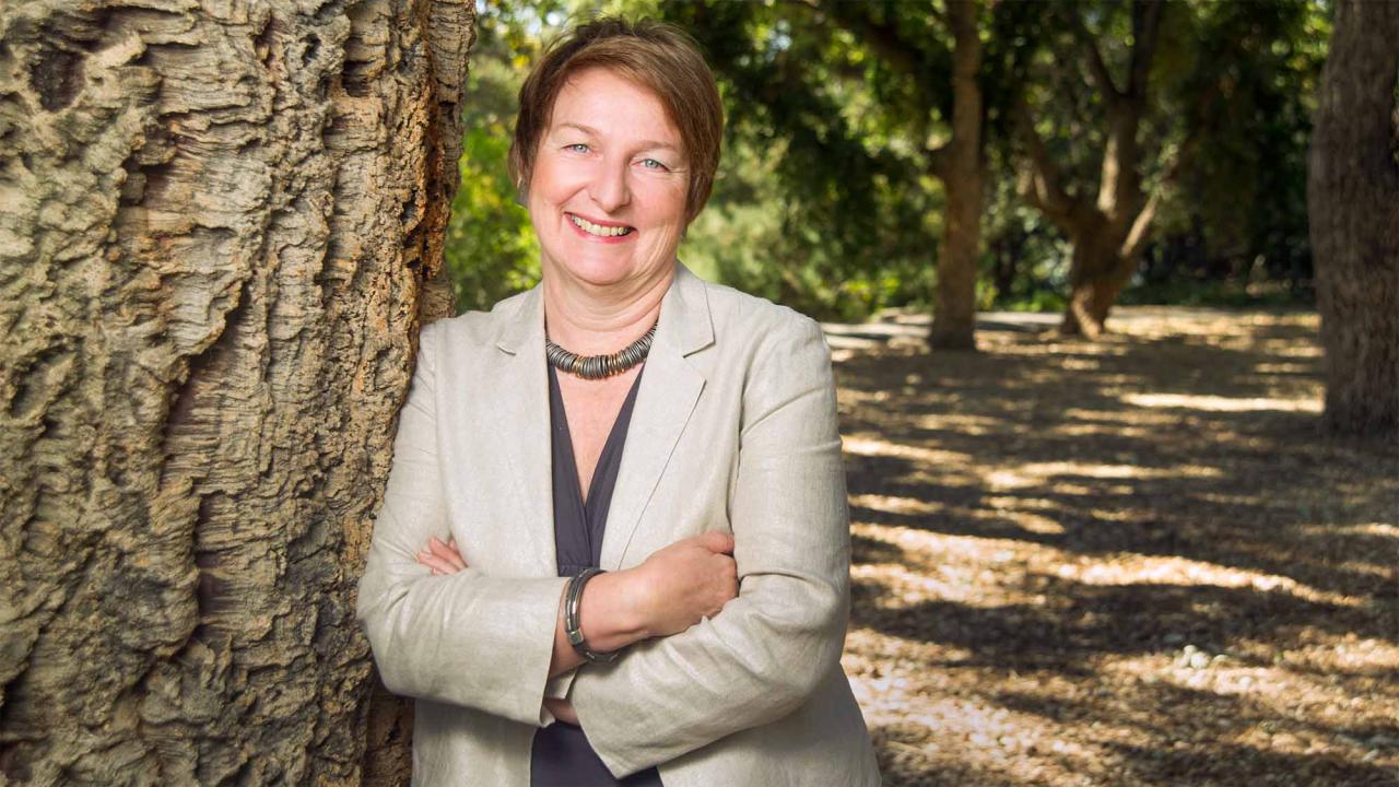 Smiling woman in beige blazer leaning against a tree in a sunlit park, portrait photo