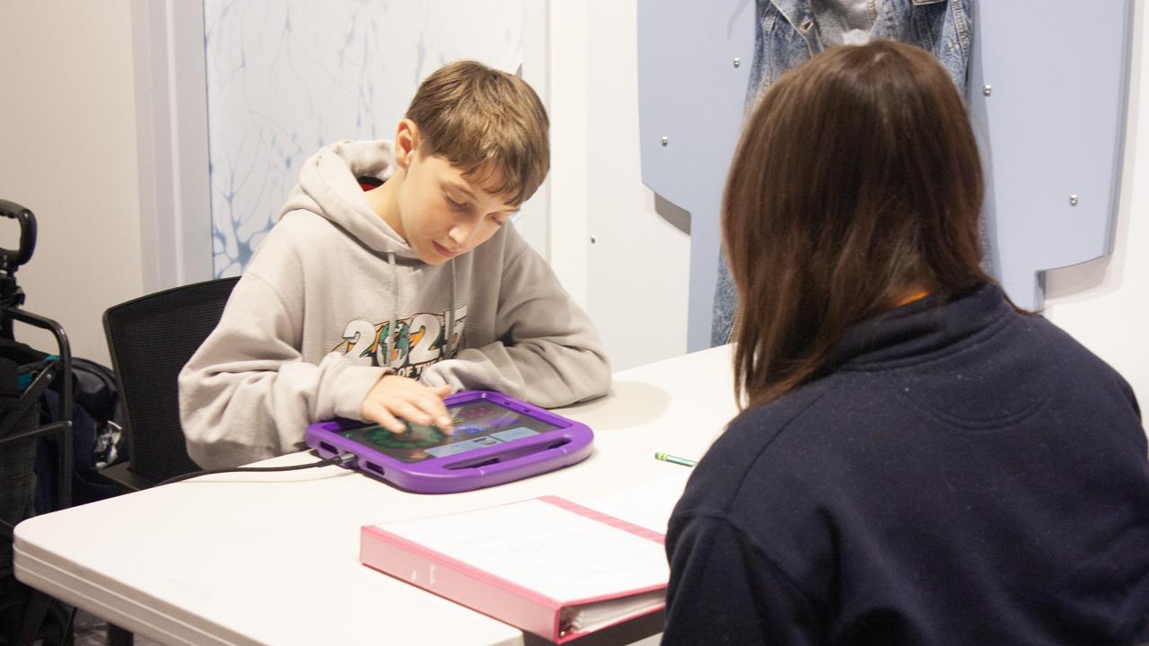 Boy focused on tablet at table, adult seated opposite observing.