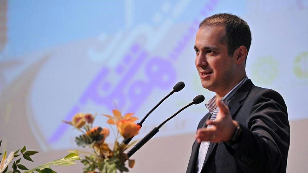 Man in suit speaking at podium with microphones, gesturing near a floral arrangement