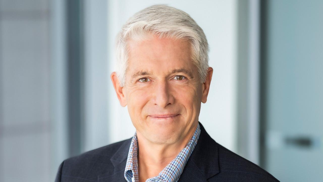 Professional headshot of gray-haired man in navy suit, slight smile, blurred office background