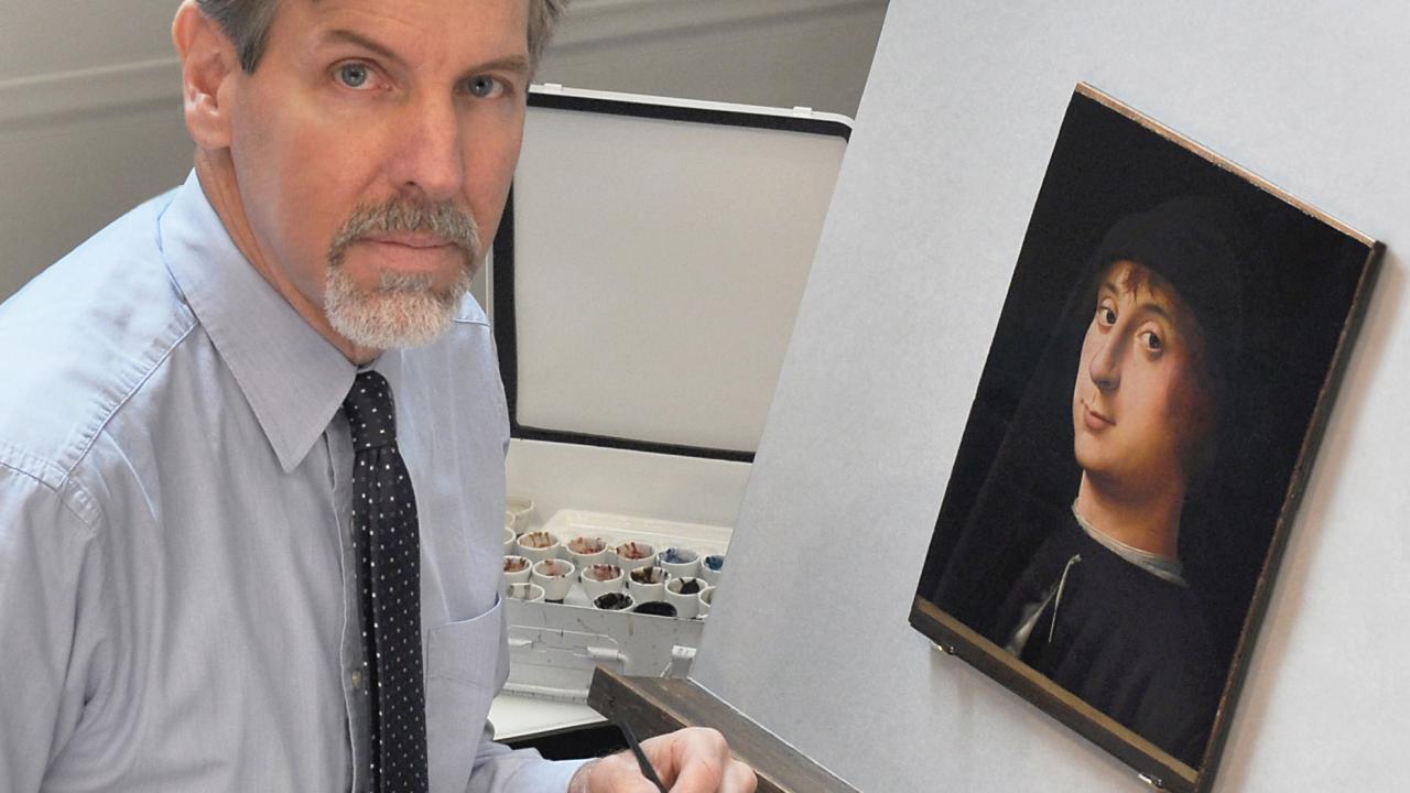 Conservator with gray hair restoring a small framed portrait on an easel