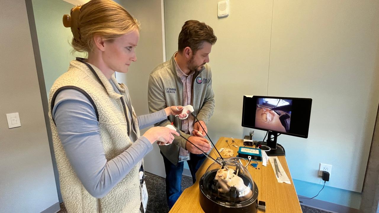 Two people work at a table with a monitor displaying a surgical procedure.