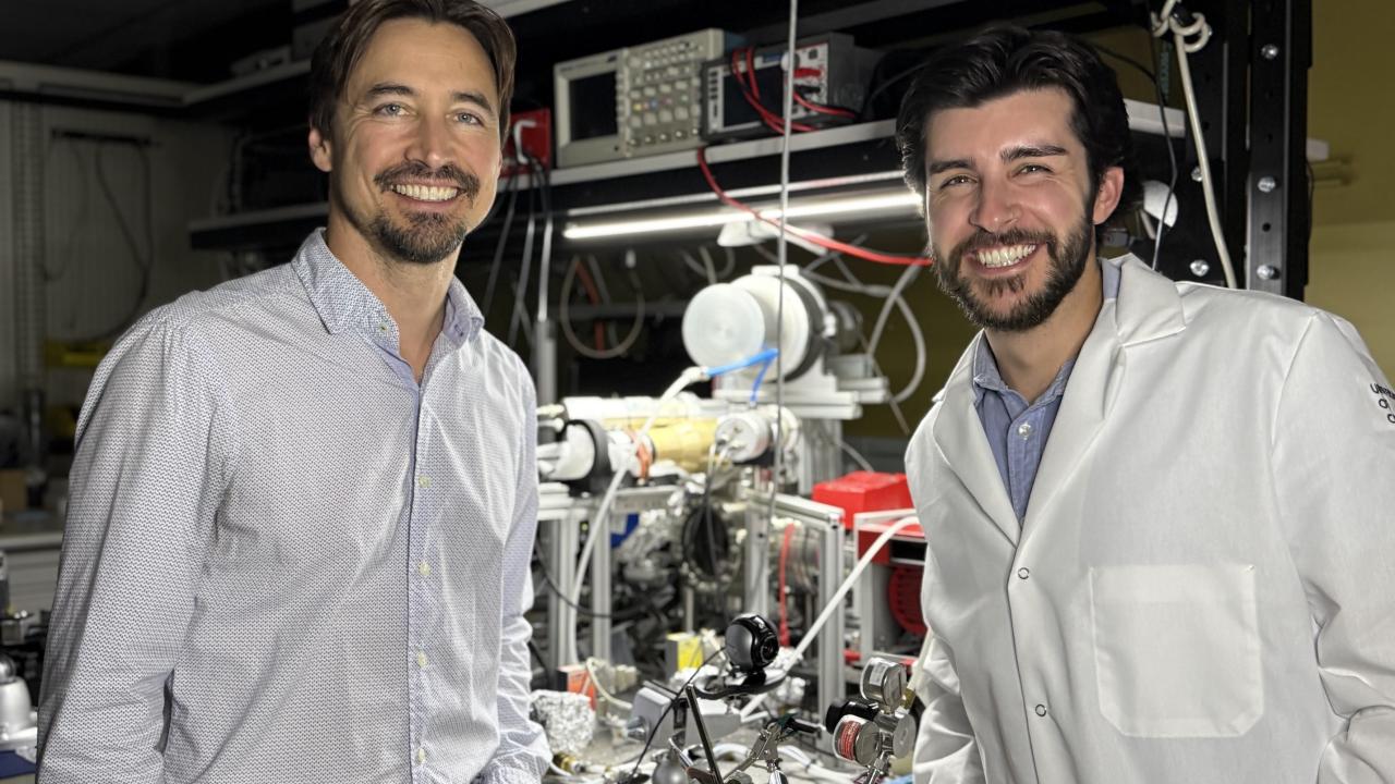 Jeremy Munday and Micah Karahadian smile beside equipmental equipmentin a lab, one wearing a white lab coat.