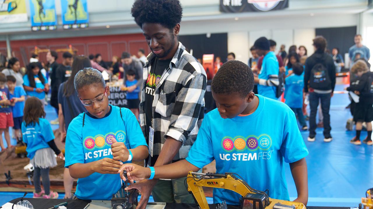 Two children in light blue UCSTEM shirts at a table with machinery. An adult in a check shirt stands behind them pointing to details. 