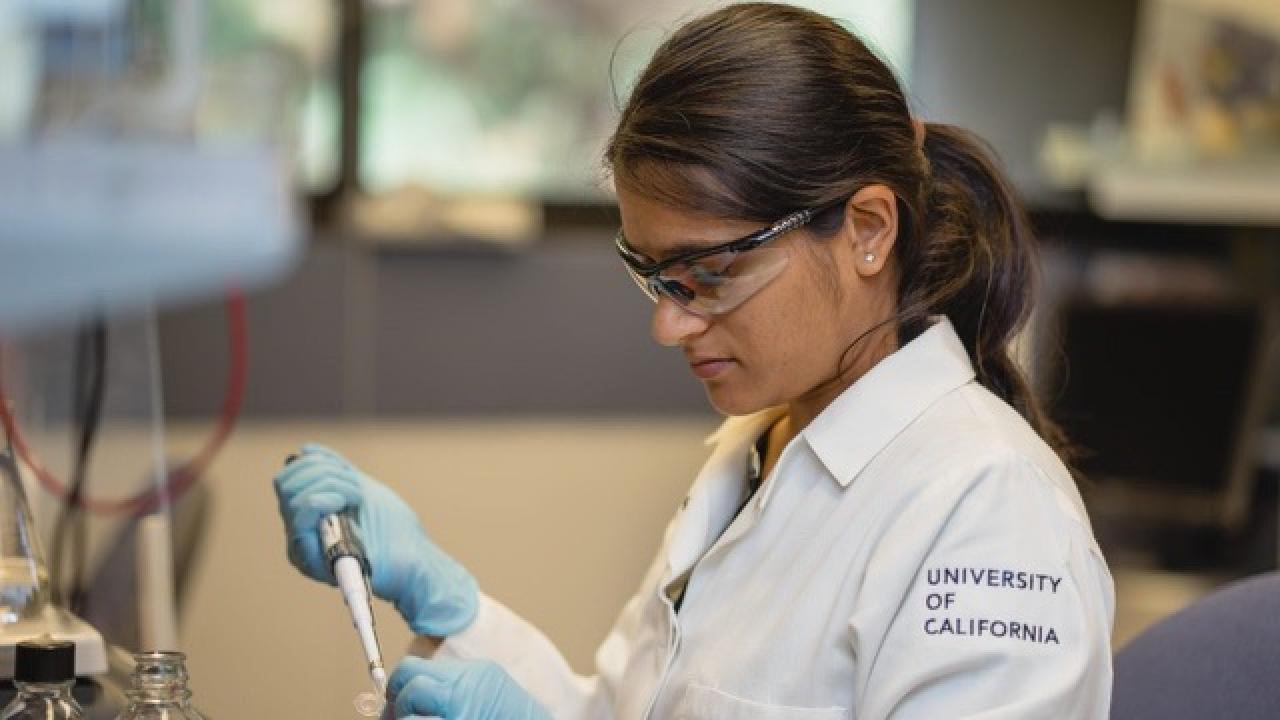 Researcher in a UC‑branded lab coat and safety gear pipetting liquid into a small container at a laboratory