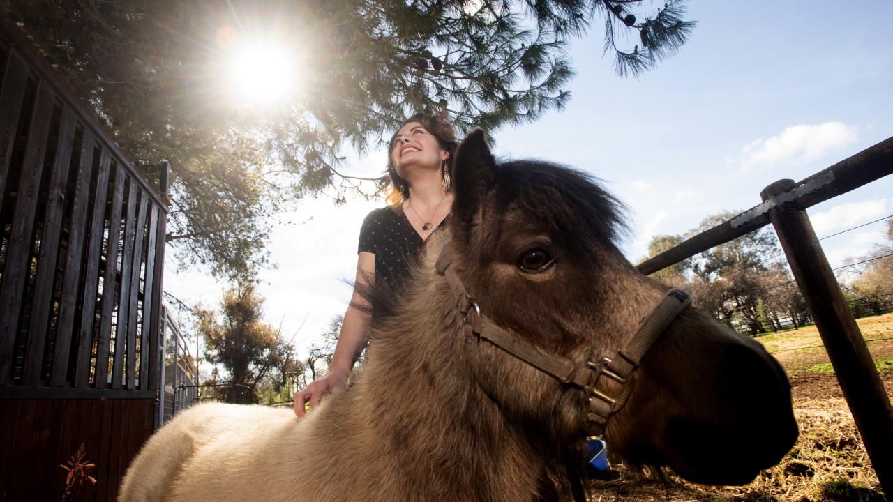 Sunlit photograph of smiling Rebecca Calisi Rodriguez standing behind a miniature horse, with trees and blue sky behind.