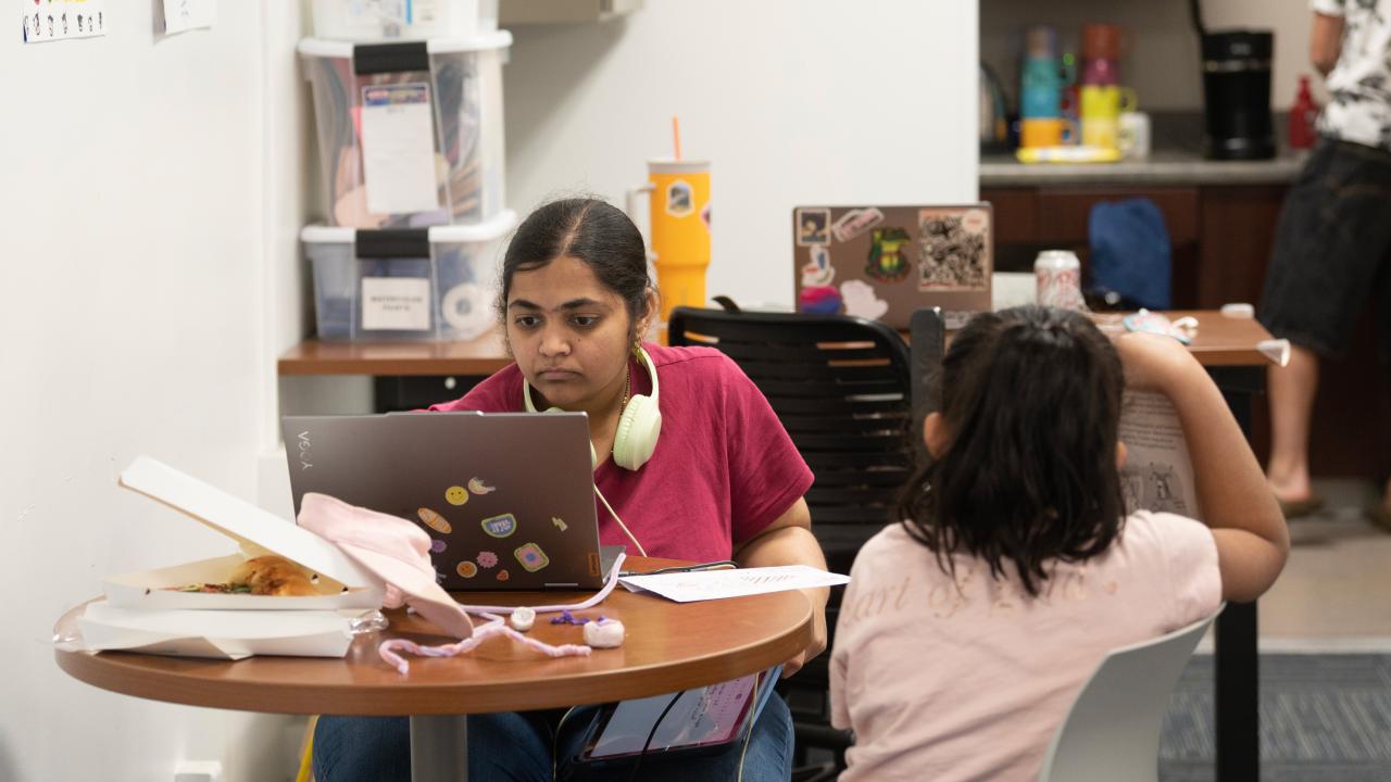 A young woman works on a laptop at a table, while a child sits nearby, looking away.
