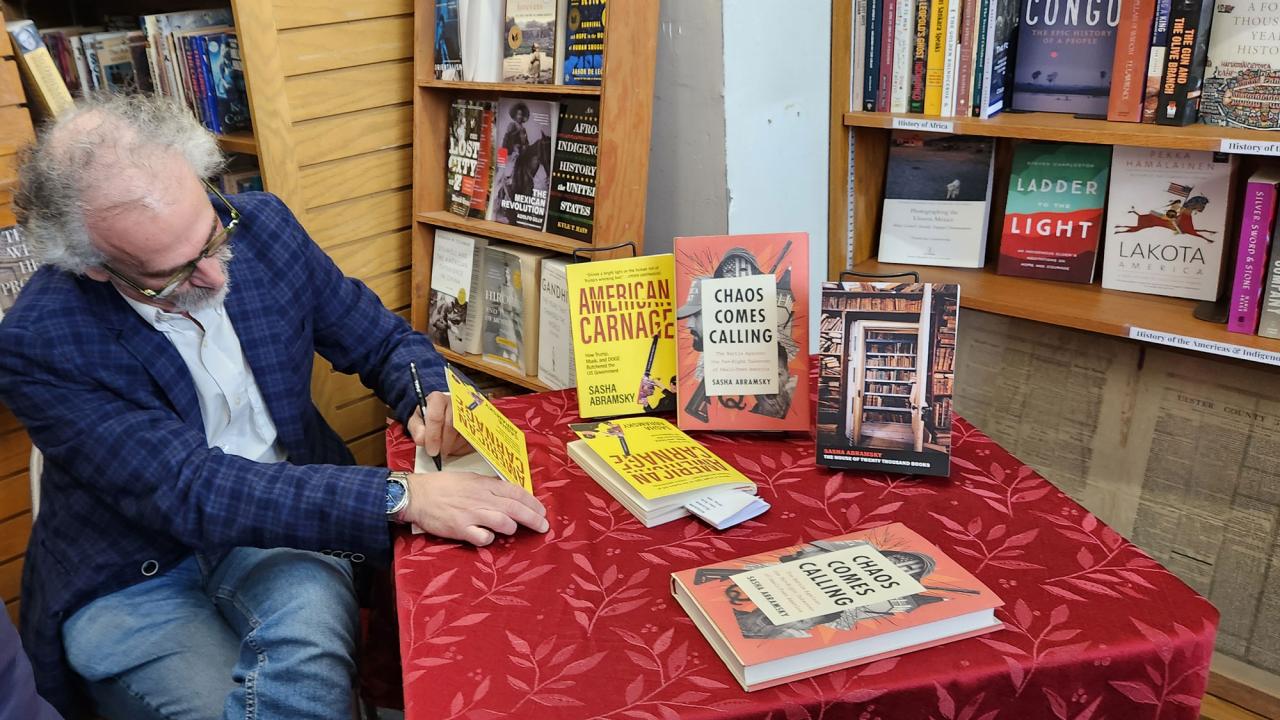 Man in blue plaid jacket signs books at bookstore table