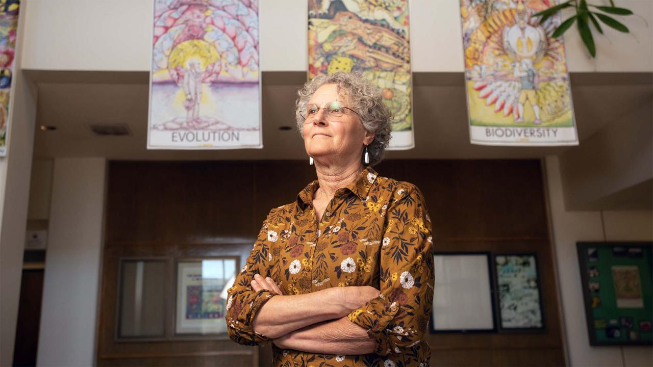 Woman in patterned blouse standing with arms crossed beneath hanging colorful art, photograph