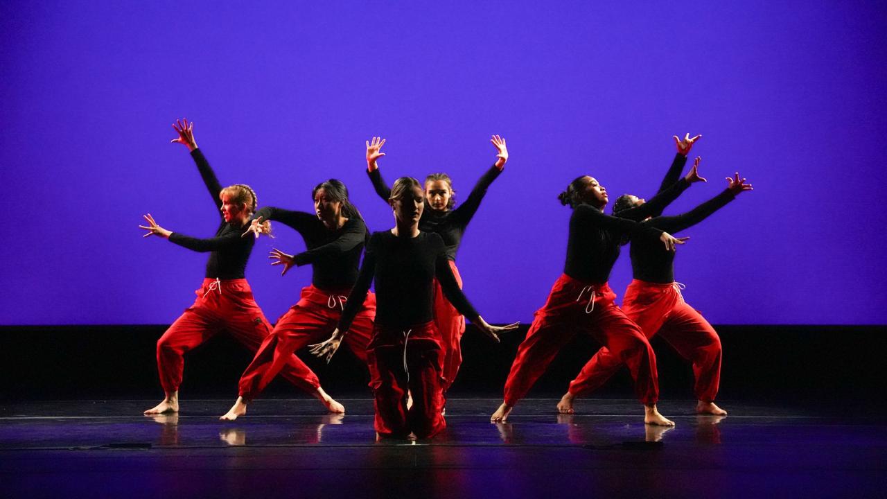 Six dancers in black tops and red pants striking energetic poses on a purple-lit stage
