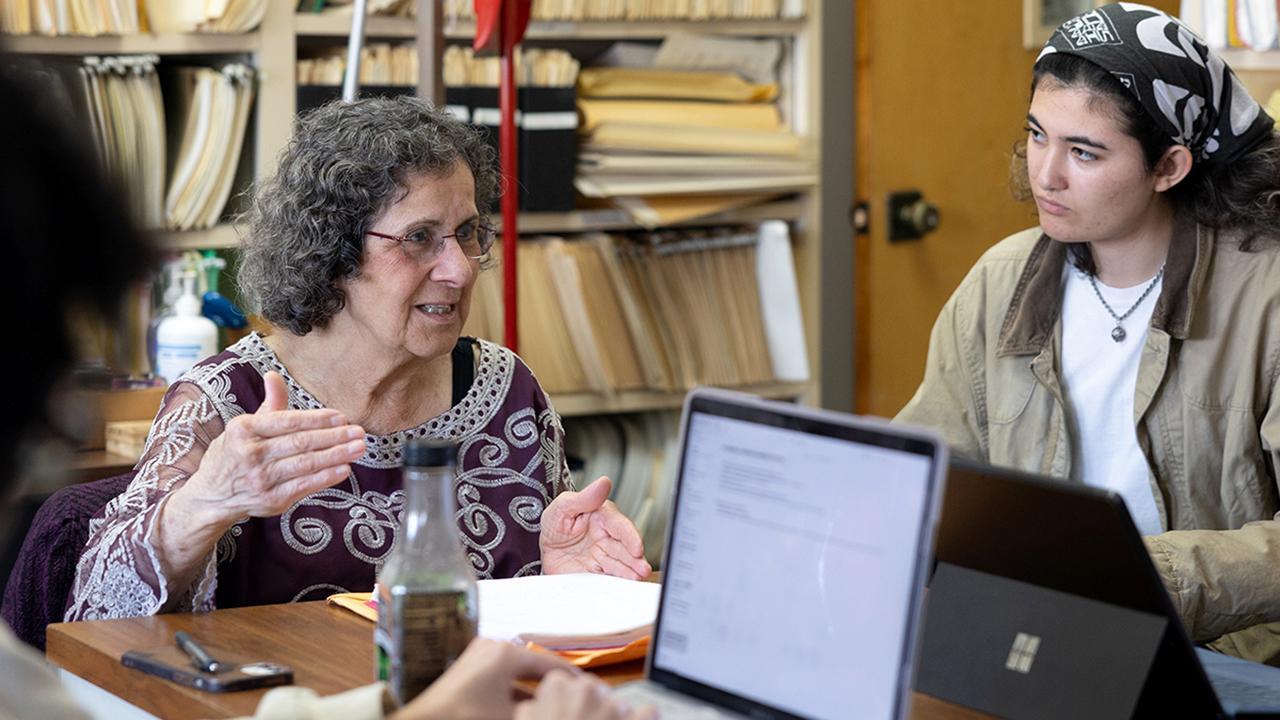 Professor of Anthropology explaining her work to a student at a table with computers and tablets.