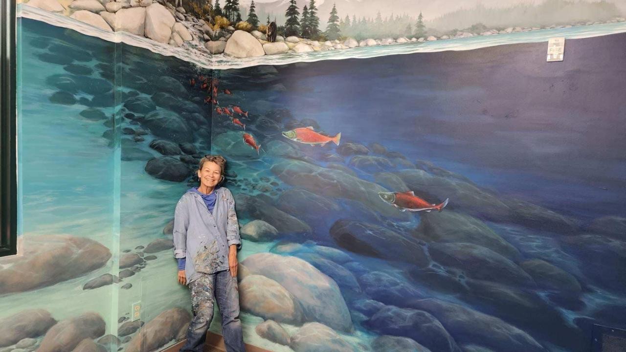 Susie Alexander stands in front of her mural, which features painted rocks, trees, and mountains, as well as rocks and fish in blue water.