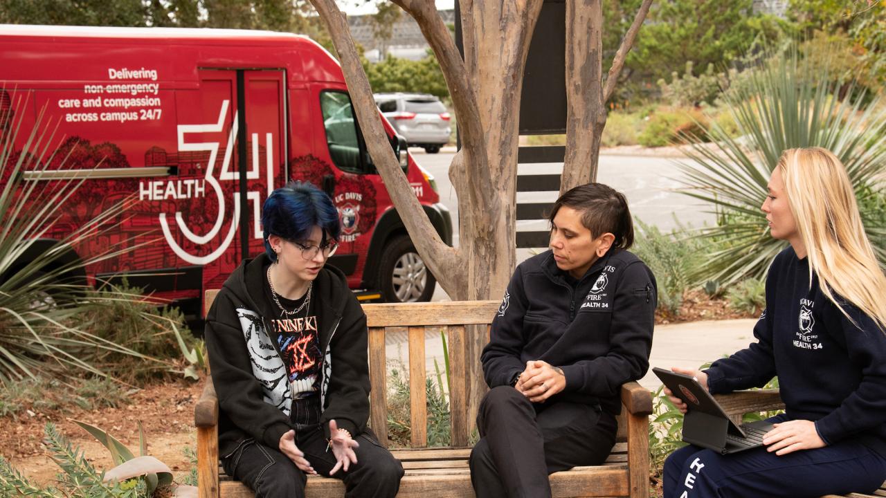 Sitting on benches, three women talk, and the Health 34 van is in the background