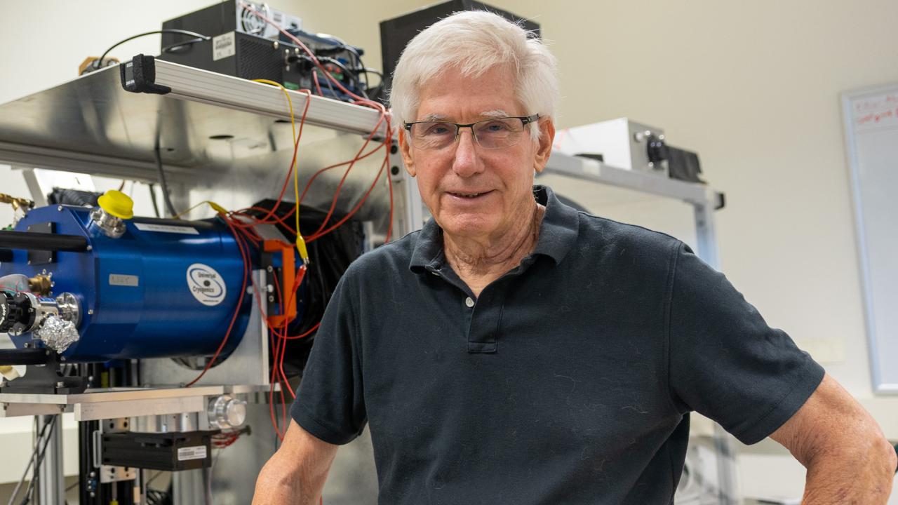 A smiling man wearing a black polo stands in front of scientific equipment.
