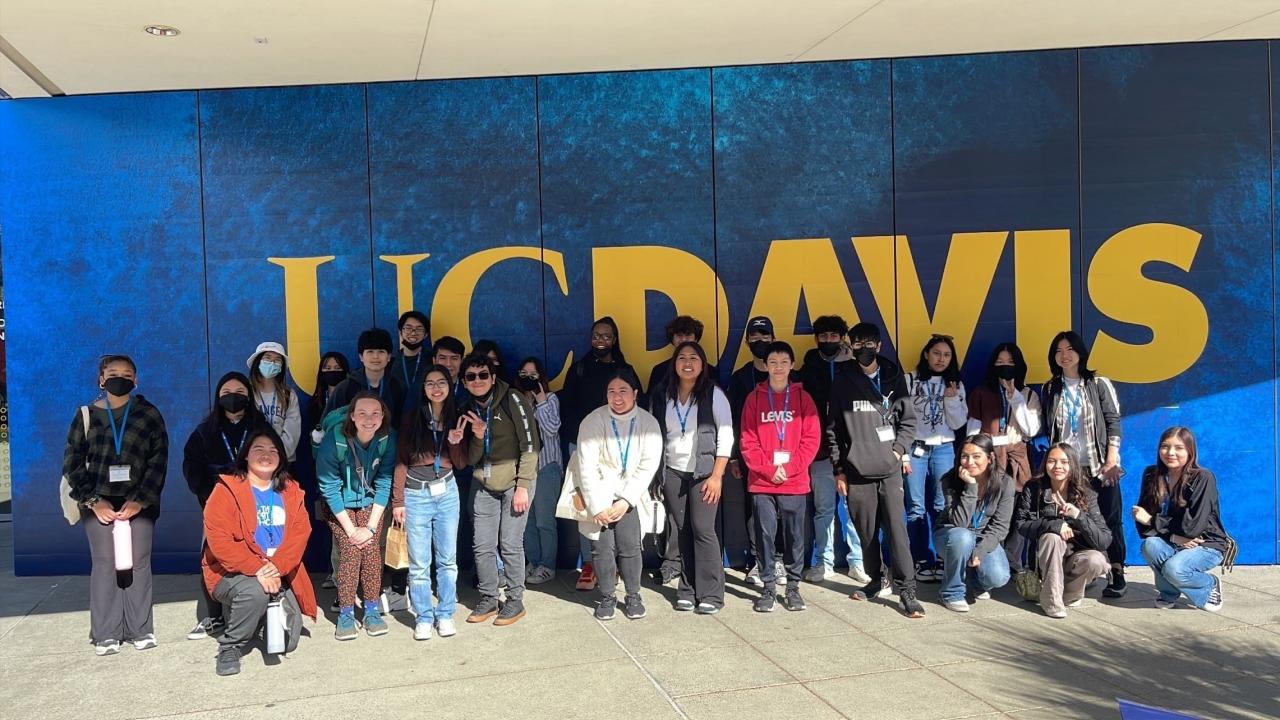 Group of students posing in front of large gold UC Davis sign on blue wall