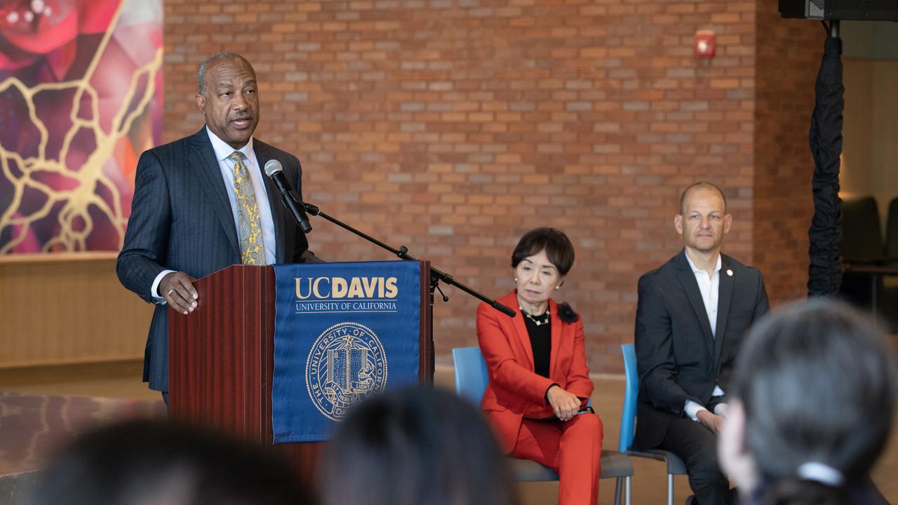 Standing at a lectern, UC Davis Chancellor Gary S. May speaks into a microphone as a seated man and woman look on