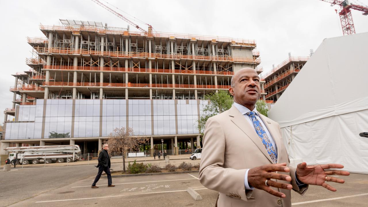 UC Davis Chancellor Gary S. May in tan suit, speaks into TV camera (not seen), while stanbding in front of Aggie Square building