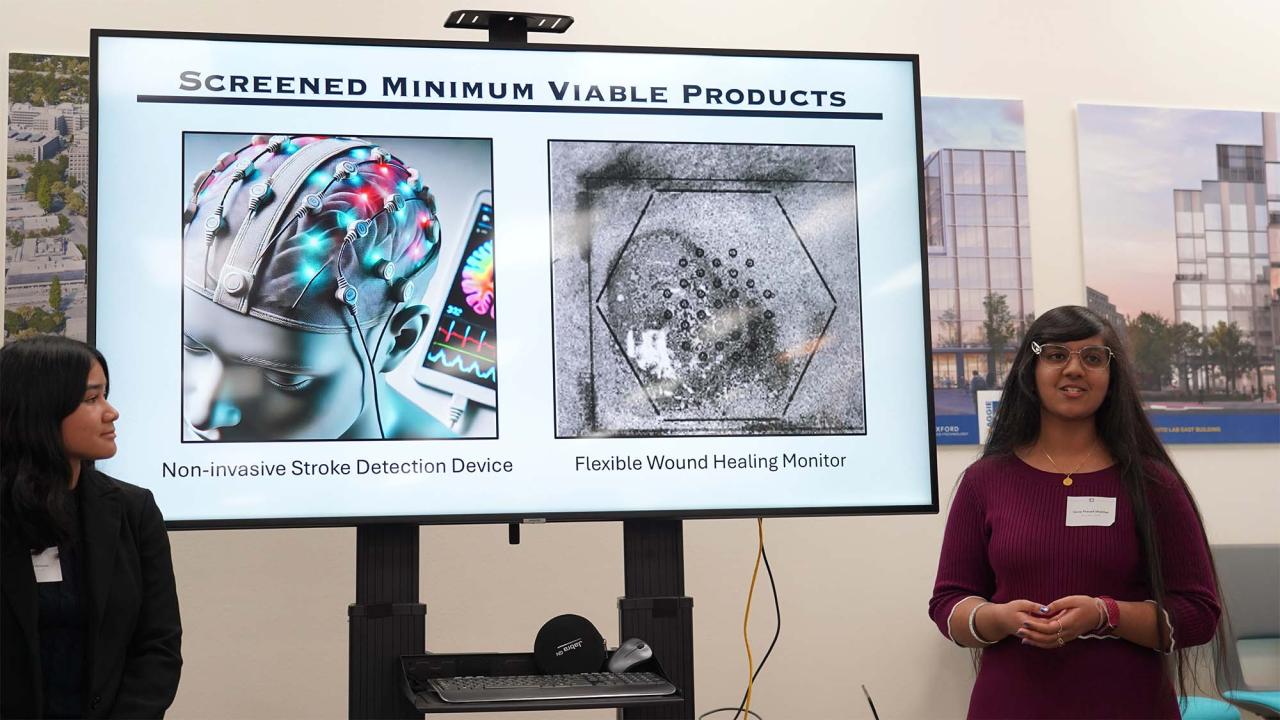 Two people stand and speak in front of a screen that reads "screened minimum viable products," showing renderings of medical devices