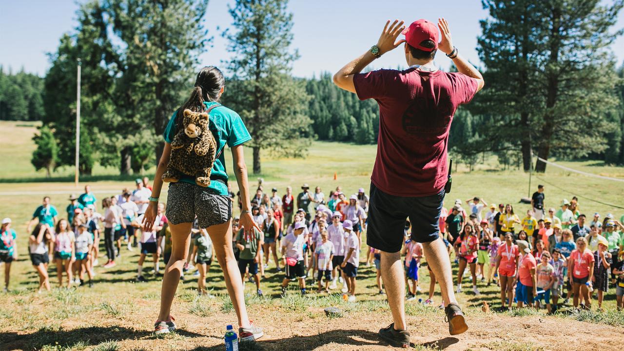 Two camp counselors stand on hill, shouting to group of campers assembled below.