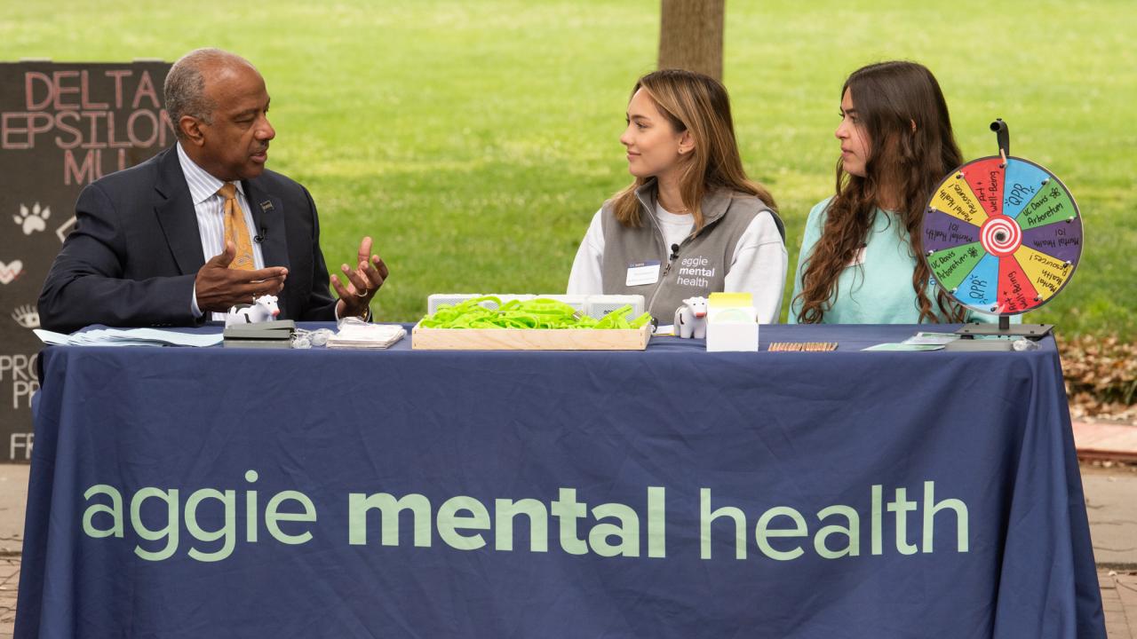 UC Davis Chancellor Gary S. May in suit, sits at table, chatting with two Aggie Mental Health Ambassadors are tabling, on the Quad