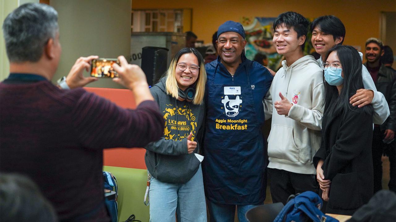 Chancellor May in "Moonlioght Breakfast" apron, flanked by students, having their picture taken