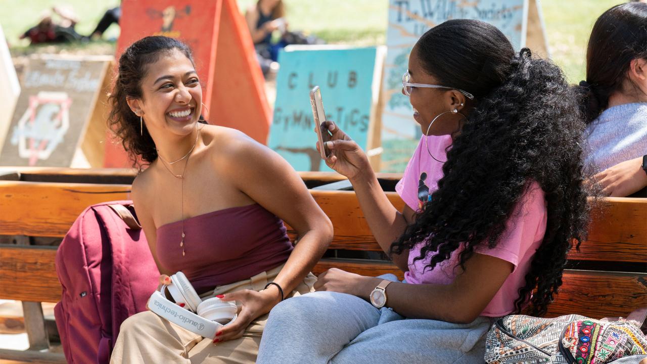 Two UC Davis students sit on a bench, smiling. The one on the right uses their phone to take a photo of the one on the left.