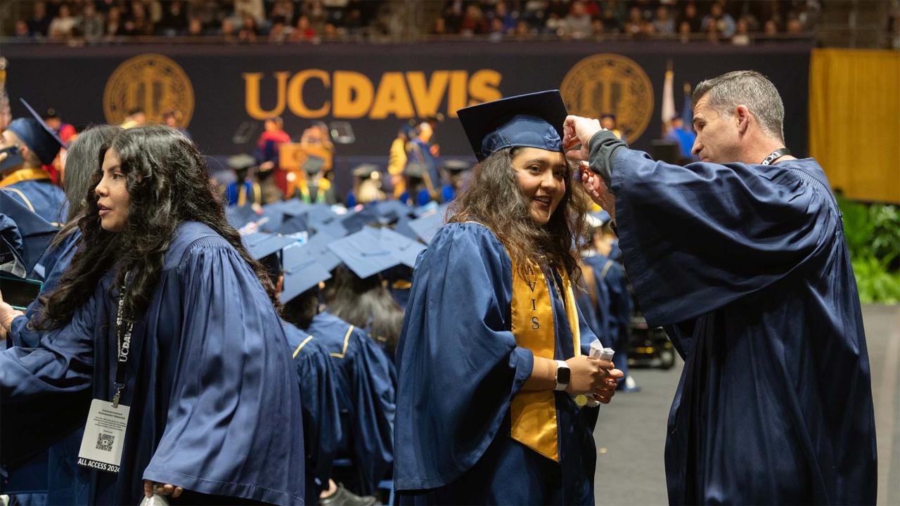 Smiling UC Davis graduate in cap and gown having tassel adjusted by staff volunteer