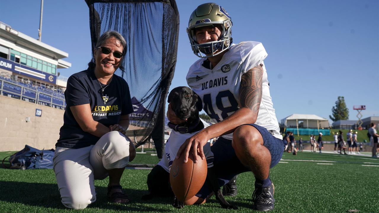 Cori, a black labrador retriever, poses for a photo with a football kicker and faculty member