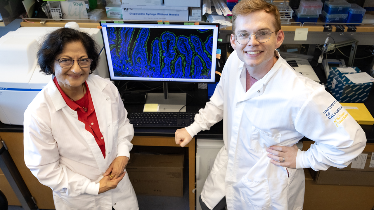 Two smiling scientists in white lab coats by a monitor showing a blue-stained microscope image