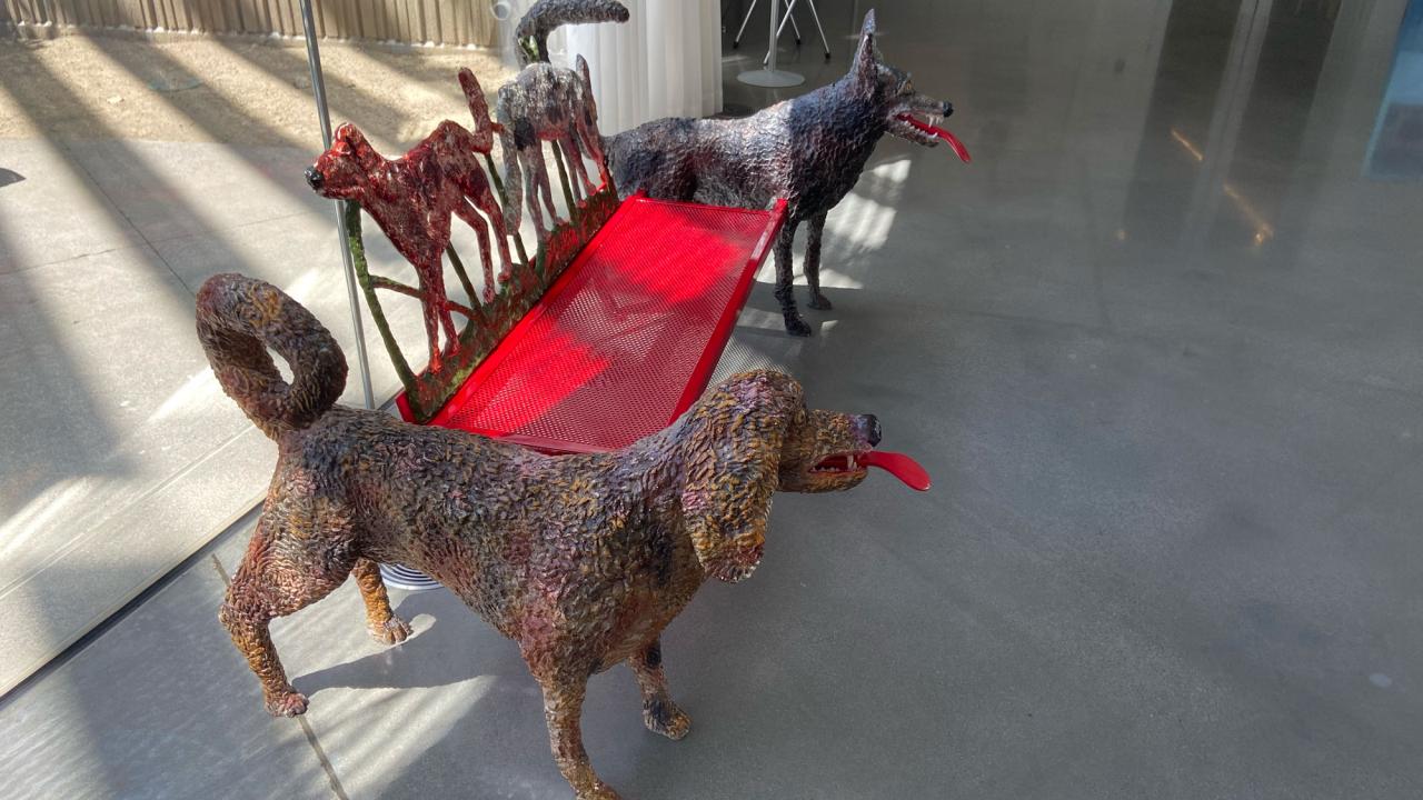 Three painted dog sculptures linked by a red bench on a sunlit concrete floor