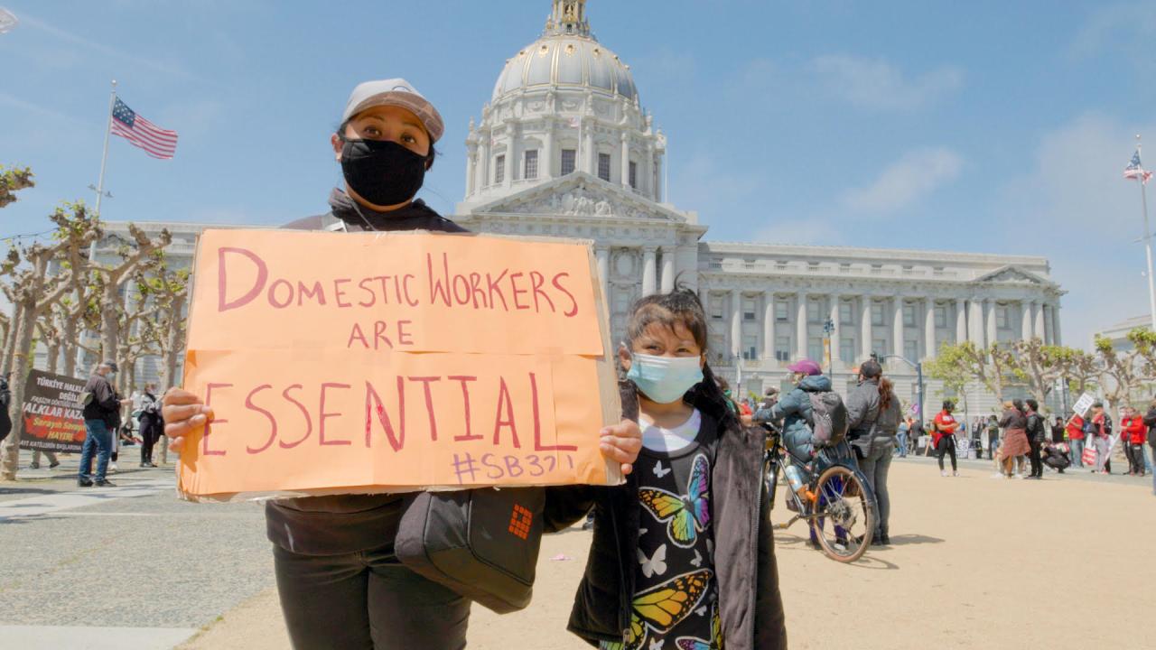 A woman and child with the San Francisco city council building in the background. Both wear facemarks. The woman holds a cardboard sign reading "Domestic Workers are Essential #SB321"