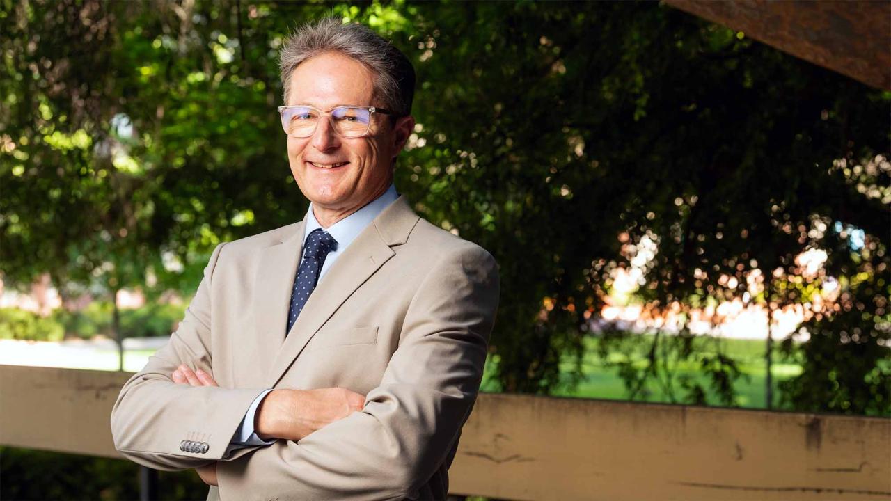 Smiling man in beige suit and glasses, arms crossed, outdoor leafy background