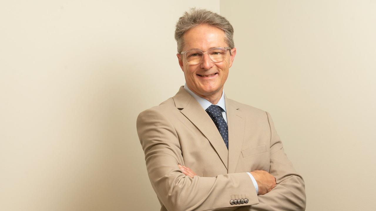 Smiling man in beige suit and blue tie, arms crossed against neutral background