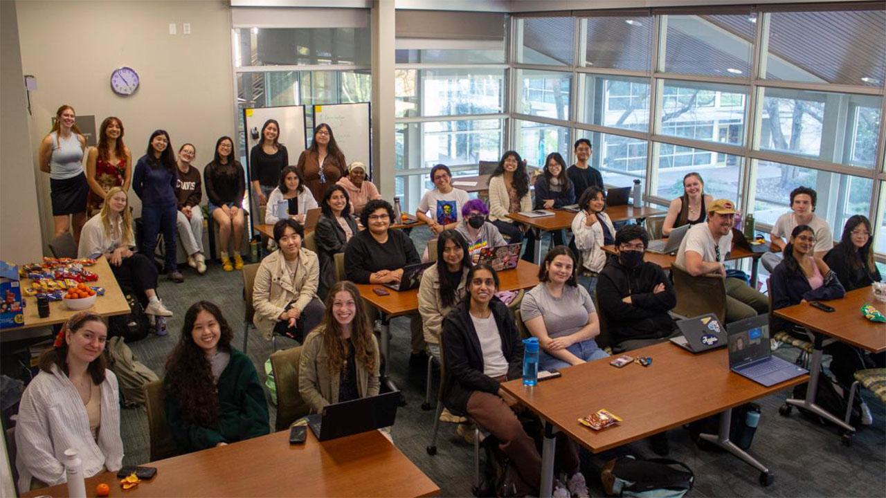 Group photo of about 30 students and instructors in a bright classroom around tables.