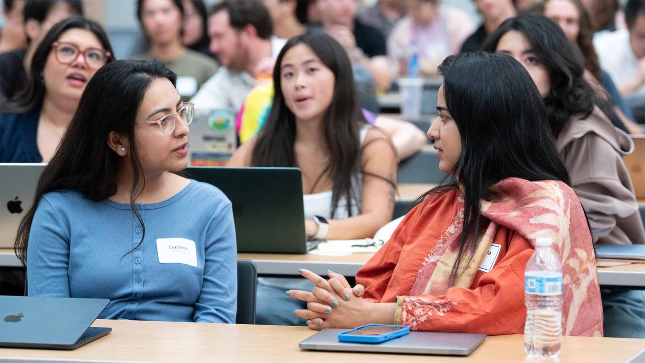 Three students conversing in a crowded lecture hall, laptops and notebooks on desks