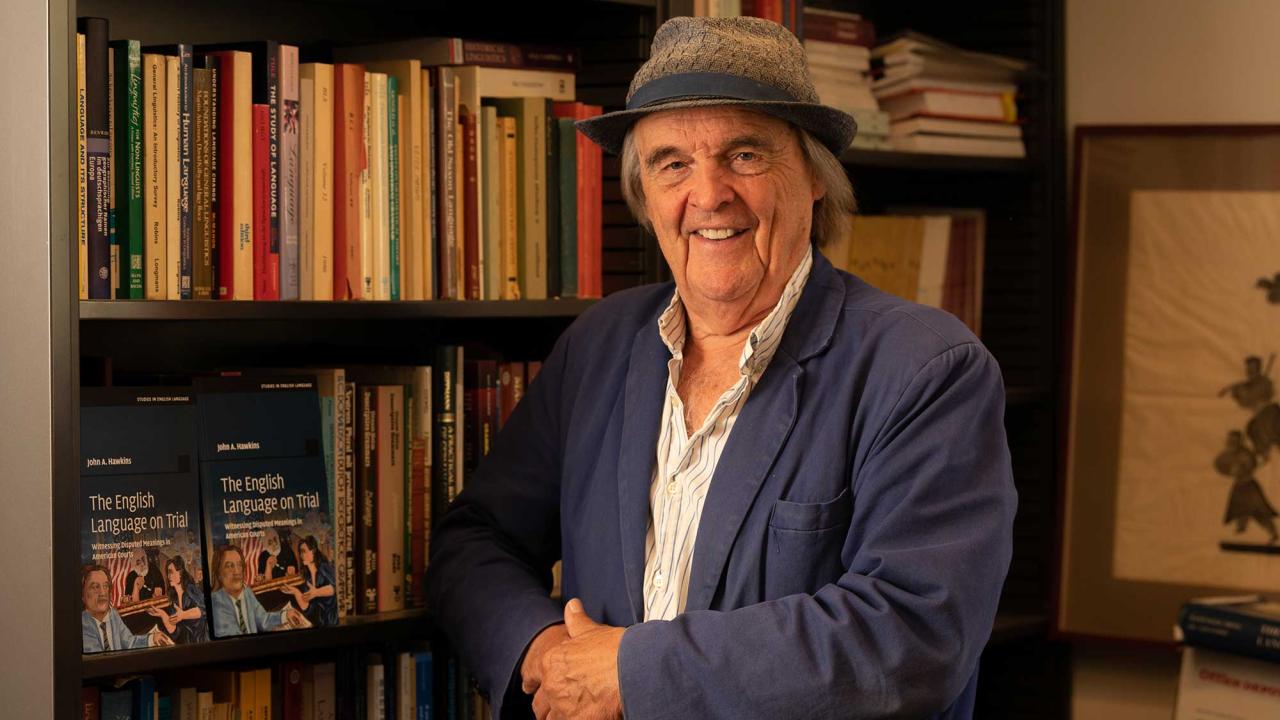 Smiling man in hat and blue jacket by bookshelf, warm-lit photograph.