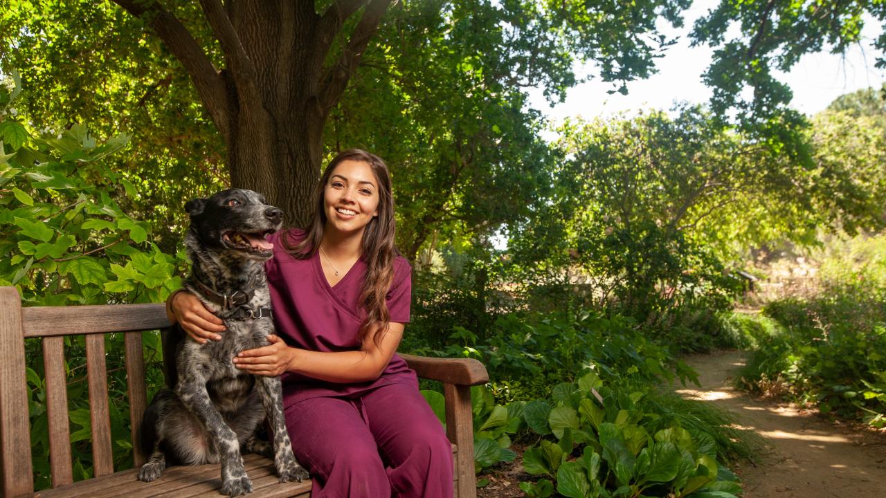 A student in scrubs poses next to a spotted dog in the leafy UC Davis Arboretum.