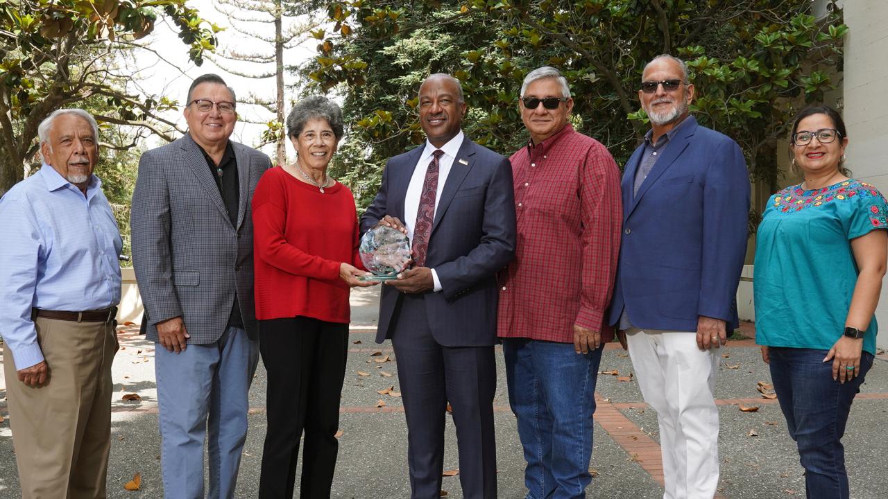 Seven people, posed in a line, with Chancellor Gary S. May and a woman holding trophy