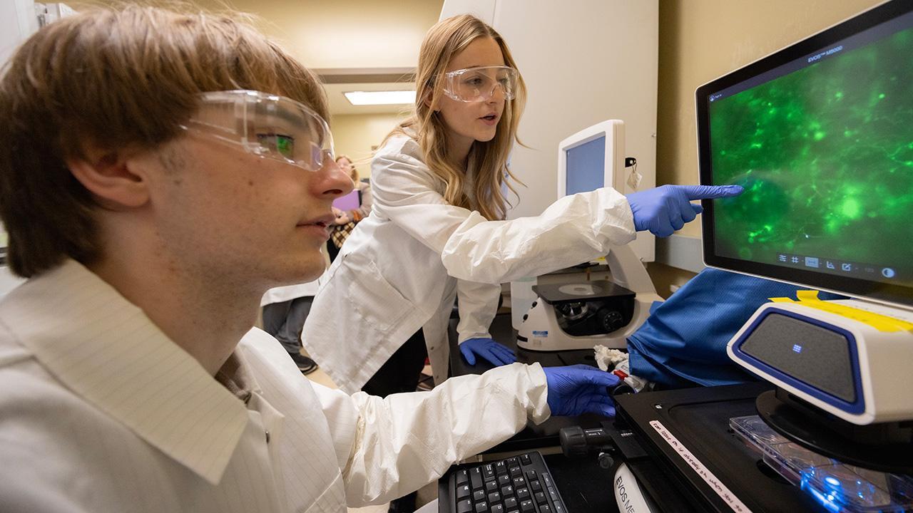 Two research students in lab coats and safety goggles examine a green display on a computer screen.