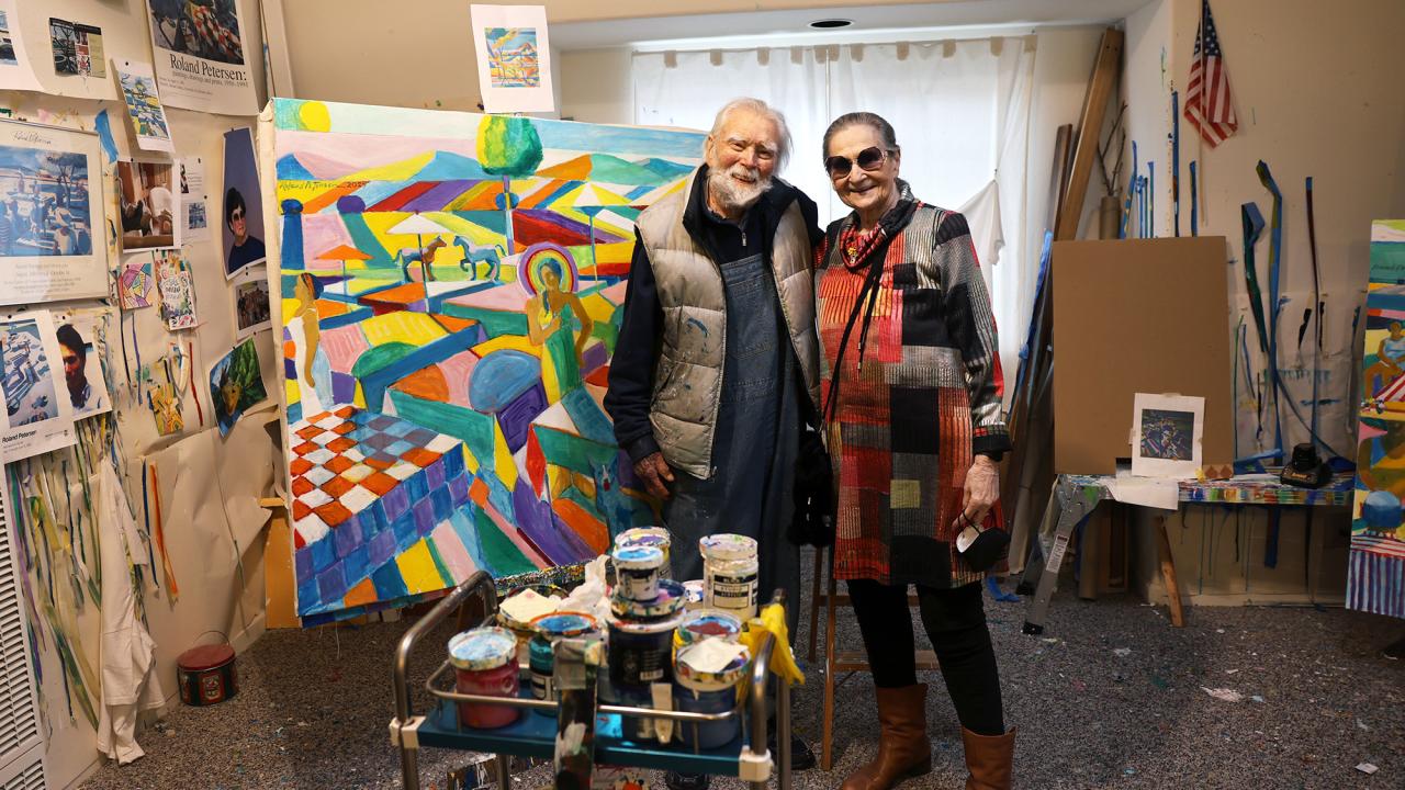 Two people pose in an art studio with a colorful abstract painting in the background.