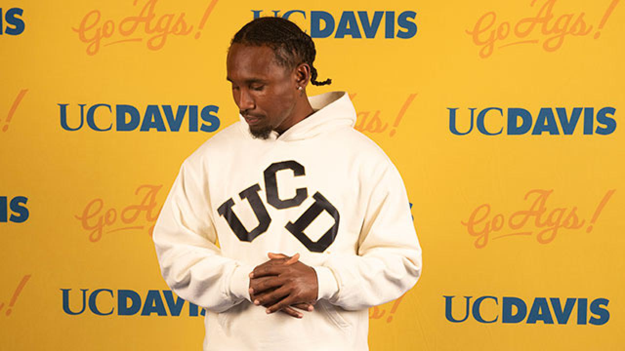 Young man in white "UCD" hoodie standing before yellow UC Davis logo backdrop