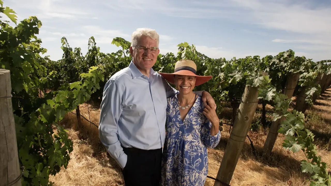Portrait of a couple standing in a vineyard on a sunny day. The man wears a light colored shirt and glasses. The woman wears a sundress and large-brimmed straw hat.