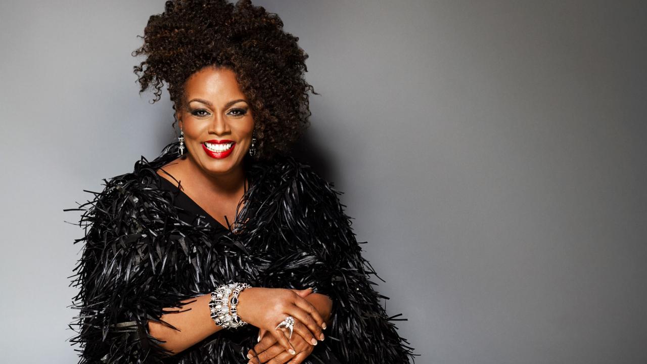 Smiling woman with curly hair wearing a black feathered coat and sparkling bracelets, gray backdrop