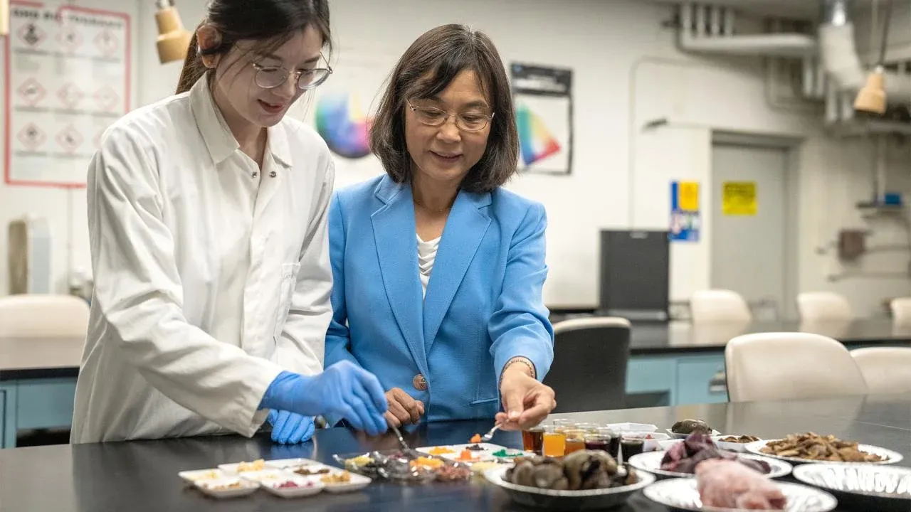 In a laboratory setting, an agricultural engineering student in a white lab coat works with their Professor, to their left, in a blue coat. They both are smiling and enjoying working together.