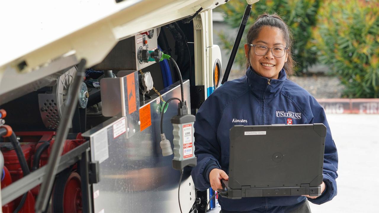 A woman holds a computer outside a bus's rear panel that is open