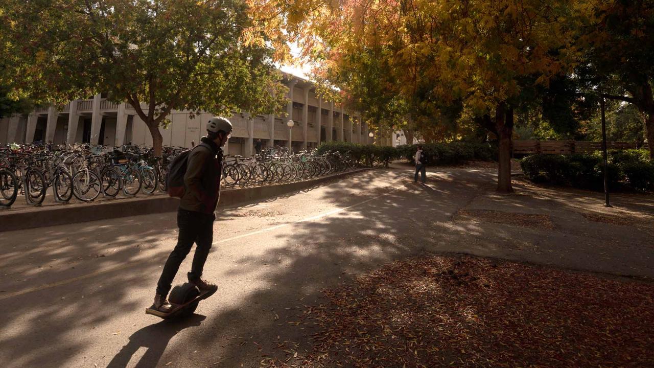 Person rides one-wheeled scooter near fall leaves.