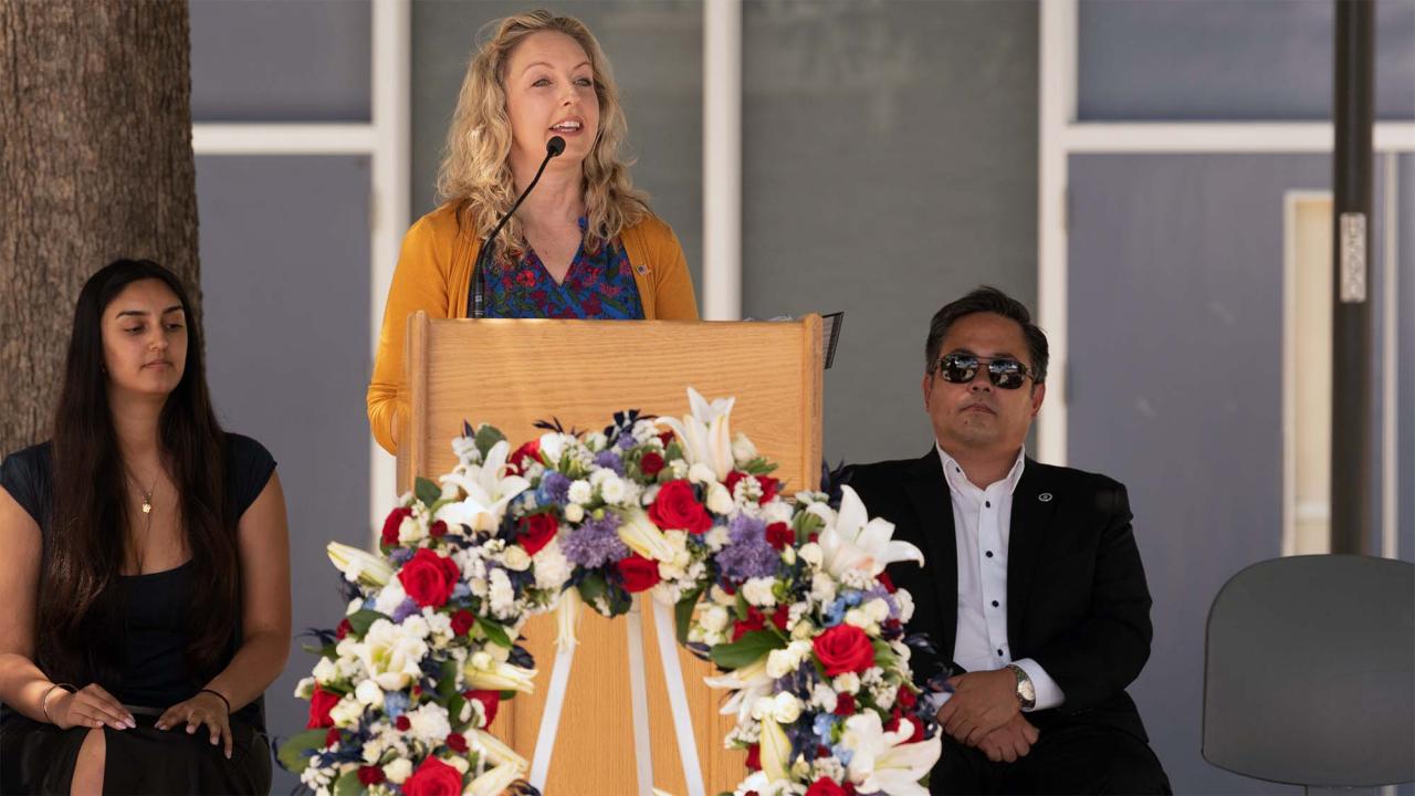 Woman speaking at podium adorned with red, white, purple floral wreath; two seated attendants
