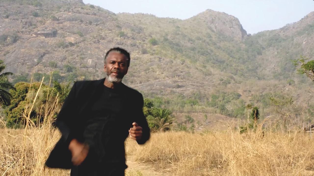 Photo of a man with brown skin in a dark suit looking at camera. He is standing in a field of tall golden grass and there are low mountains in the background.