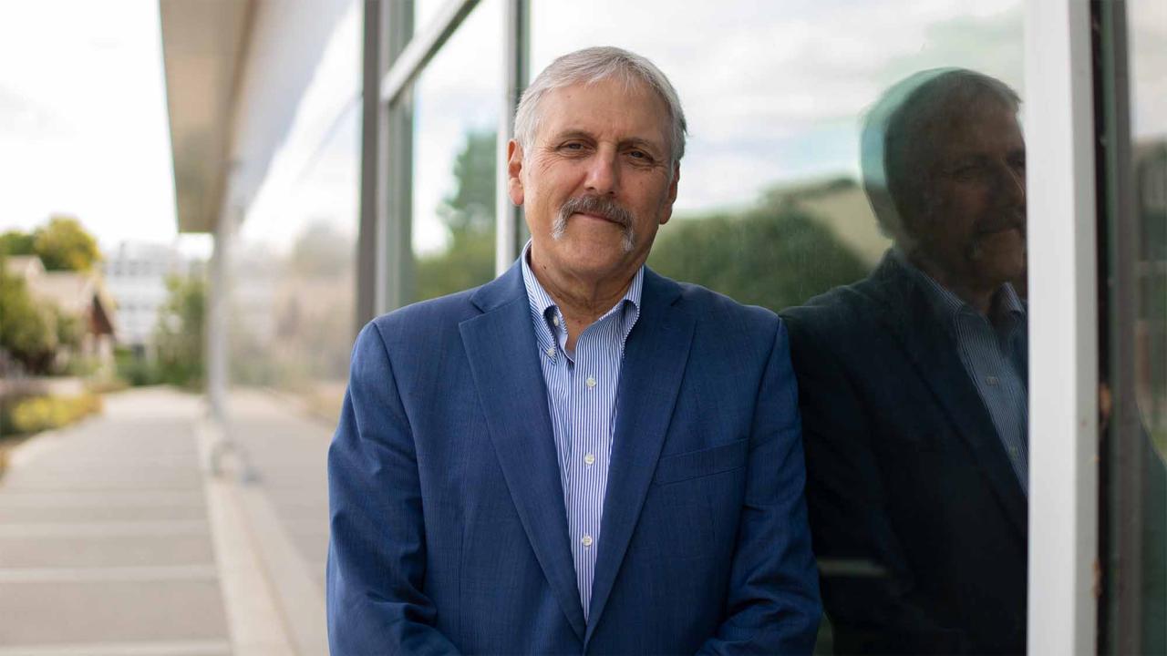 Man with gray hair and mustache in blue suit leaning against glass wall