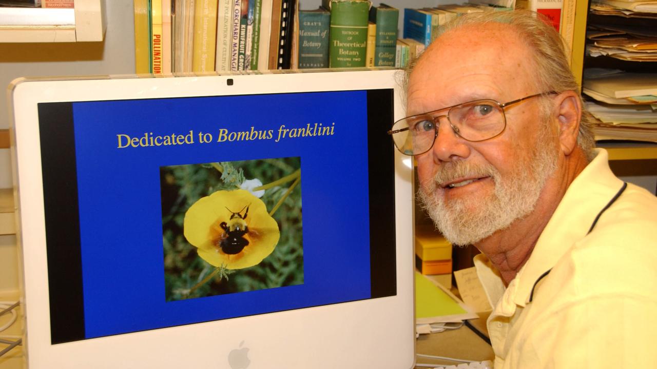An older man with thinning hair and glasses poses next to a computer screen. On screen is a photo of a bee in the middle of a flower with labeliing "Dedicated to Bombus franklini"